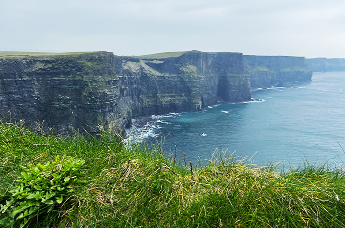 A panoramic view of ocean waves crashing against the Cliffs of Moher, seen from a lush grassy hilltop.