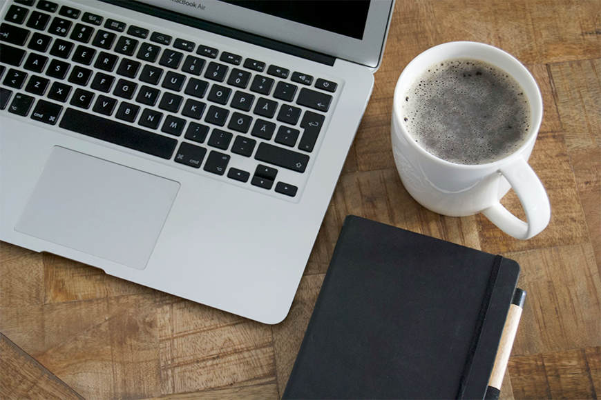Photo of an open laptop computer on a wooden desk beside a cup of coffee and notebook.