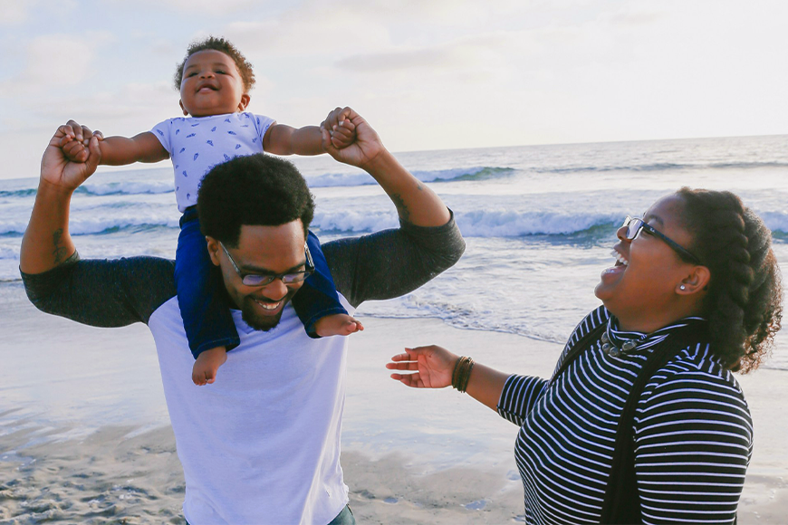 A happy African- American family smiling on a beach while the father holds his son on his shoulders and the mother looks on with laughter. They are not stressed out about their trip because they planned in advance with My Tailored Escape, a boutique travel agency.