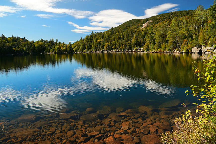 Clear alpine lake reflecting autumn foliage and blue sky in the Adirondacks along the scenic Adirondack train route, one of the best places to visit for fall foliage, by My Tailored Escape, boutique travel agency serving Washington DC, Maryland, Virginia and nationwide.
