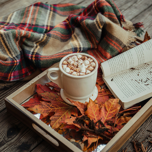 Cozy autumn scene with hot cocoa, a plaid blanket, an open book, and red fall leaves on a rustic tray, illustrating the best places to travel to in the fall, by My Tailored Escape, boutique travel agency serving Washington DC, Maryland, Virginia and nationwide.