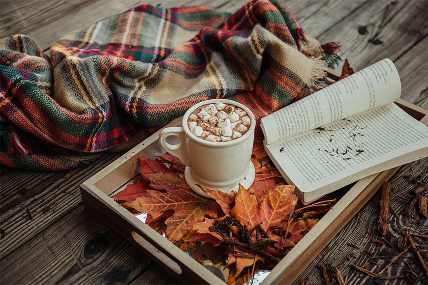 Cozy autumn scene with hot cocoa, a plaid blanket, an open book, and red fall leaves on a rustic tray, illustrating the best places to travel to in the fall, by My Tailored Escape, boutique travel agency serving Washington DC, Maryland, Virginia and nationwide.