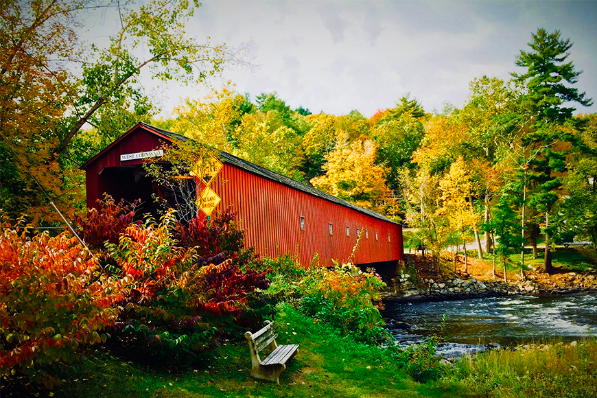 Classic red covered bridge over a rushing stream framed by red, gold, and green fall foliage in New England, one of the best places to visit for fall foliage in the US, by My Tailored Escape, boutique travel agency serving Washington DC, Maryland, Virginia and nationwide.
