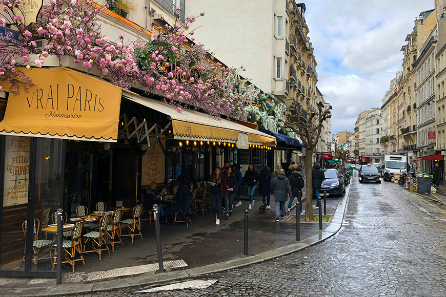Charming Montmartre café terrace with pink blooms and rattan chairs on a rainy Paris street in fall, one of the best places to visit in the fall, by My Tailored Escape, boutique travel agency serving Washington DC, Maryland, Virginia and nationwide.